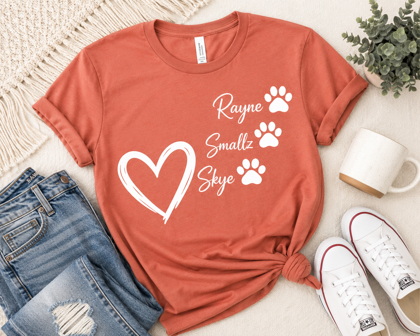 A terracotta orange t-shirt with a white graphic print reading 'Rayne Smallz Skye' and paw prints, styled in a flat lay with blue jeans, white sneakers, a mug, and a plant 
