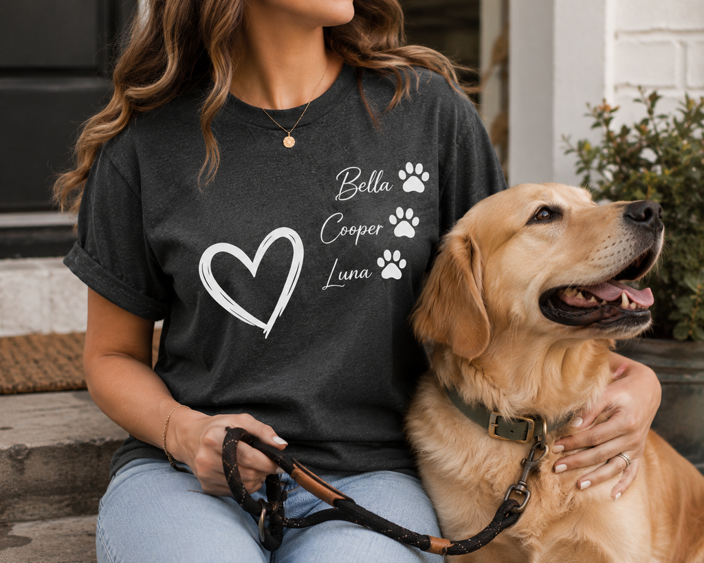 A woman wearing a dark charcoal gray t-shirt with a white graphic print of a heart and the names 'Bella', 'Cooper', and 'Luna' alongside paw prints, sitting with a golden retriever dog on a porch.